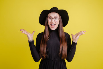 Surprised girl in a black hat and glasses spread her hands to the sides in an isolated room on a yellow background