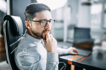 pensive bearded man with glasses on the workplace in the office reflects. The concept of a difficult task at work