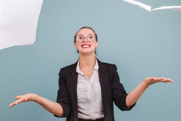 Young office worker girl in glasses in classic suit threw up clean sheets of paper isolated indoors on blue background looking at camera