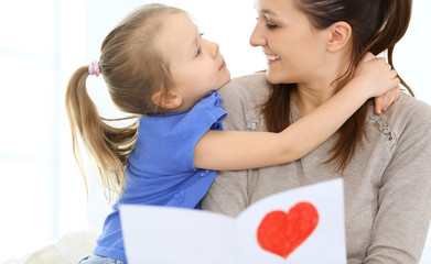 Mother's day concept. Child daughter congratulates mom and gives her postcard with red heart shape. Mum and girl happy smiling and hugging. Family fun and holiday 
