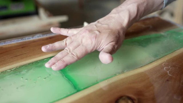 Close-up hands of mature furniture maker creating epoxy resin decoration on unfinished wooden tabletop and touching smooth surface