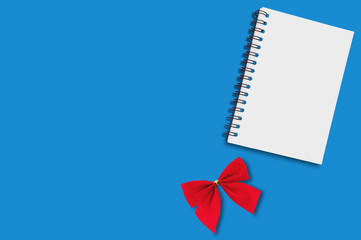 One blank paper notebook with spiral wire and red festive textile tied bow on blue table. Copy space for your text. Top view. Holidays concept