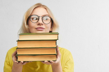 Funny clever girl looks out of a pile of books. Blonde nerd in round glasses on a white background. Educational concept