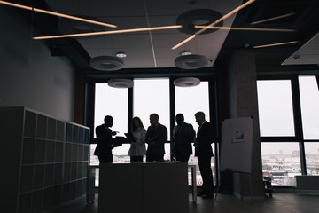 Corporate business team working in the office space standing at the table. Dark photo silhouettes of businessmen on the background of the window