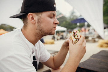 Stylish hipster man eating delicious vegan burger at street food festival. Hungry man biting burger with vegetables in summer street.