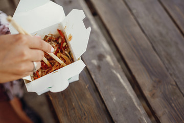 Girl eating wok noodles with vegetables and seafood in carton box to go, with bamboo chopsticks, closeup. Traditional Asian cuisine at Street food festival. Thai noodles in paper box takeaway