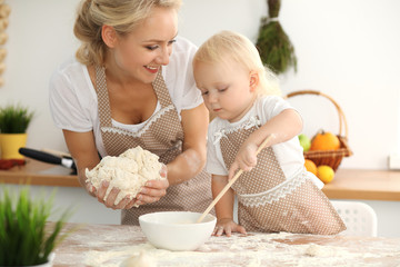 Little girl and her blonde mom in beige aprons  playing and laughing while kneading the dough in kitchen. Homemade pastry for bread, pizza or bake cookies. Family fun and cooking concept