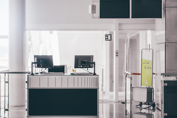 An empty gate of a modern airport terminal with computer monitors on the counter, information screens on the top, baggage gauge, and the bright corridor leading to the airplane to boarding