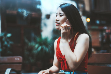 A cheerful charming Asian woman in red shirt and dress is talking on the phone with someone and smiling while sitting outdoors on a wooden bench with a building facade in a defocused background