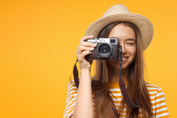 Close-up studio portrait of happy smiling young female tourist holding camera, isolated on yellow...