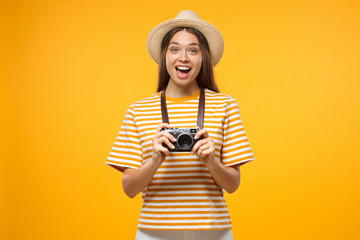 Excited young cheerful female tourist holding photo camera, isolated on yellow background.
