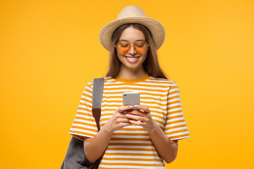 Studio portrait of happy smiling young female tourist holding phone, isolated on yellow background