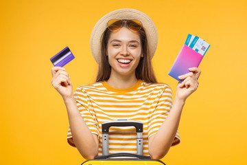 Young laughing female tourist holding credit card and passport with tickets, isolated on yellow...