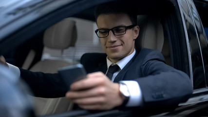 Glad businessman texting with girlfriend and smiling, sitting in car, smartphone