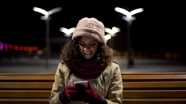 Biracial girl sitting outdoors, watching funny images and sincerely smiling