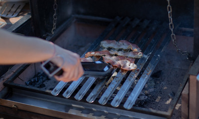 Bacon cooking on the charcoal barbecue with hands holding a pair of tongs