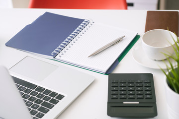 Closeup of white desktop with laptop, coffee cup, notepads, pen, calculator and other items.