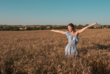 Naklejka na ściany i meble A beautiful and young girl is standing in a summer wheat field. Arms outstretched happy smiles rejoices sunrise and sunset. Free space for text. Emotions of happiness tenderness in a blue dress.