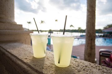 Two delicious glasses of lemonade poolside at a tropical outdoor resort. Selective focus on the two glasses on a hot summer day - obrazy, fototapety, plakaty