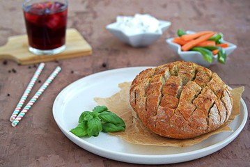 Party set on a brown concrete background: baked party bread with herbs and olive oil, a dip of cream cheese with vegetables, sliced raw carrots and green sweet pepper, grape juice with ice, straw