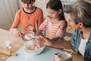 Grandmother putting flour with wooden spoon in sieve in kitchen
