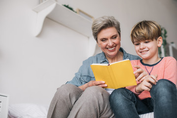 Low angle of little boy reading book with his grandmother in house