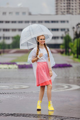 Young pretty girl with two braids in yellow boots and with transparent umbrella stands near fountain.