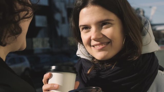 Close up portrait of two girls talking outside smiling holding coffee cup 4k. Attractive women friends having chat sharing secrets in morning street natural light. Lifestyle conversation friendship