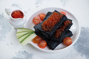 White plate with black stuffed crepes and red caviar over white concrete background, horizontal shot