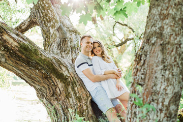 Young pretty couple in love lying on tree in park. Handsome cheerful blonde girl in white dress hugging her boyfriend. Man and woman having fun outdoors