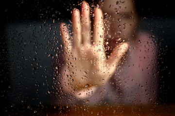 Fototapeta na wymiar Female hand palm silhouette behind the window with raindrops reaching for the glass. A request for help, depression, stress blurred bokeh background. Refusal denial of alcohol and drugs