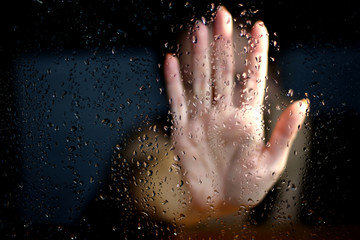 Female hand palm silhouette outside the window with raindrops reaching for the glass. A request for help, depression blurred bokeh background