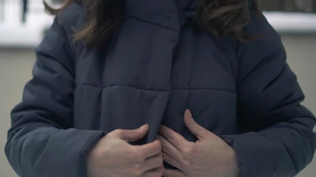 Slowmotion tilt down of young brunette woman zipping her down grey long winter coat outdoors. Female hands with a ring zipping up a long winter brand new down coat with pockets.