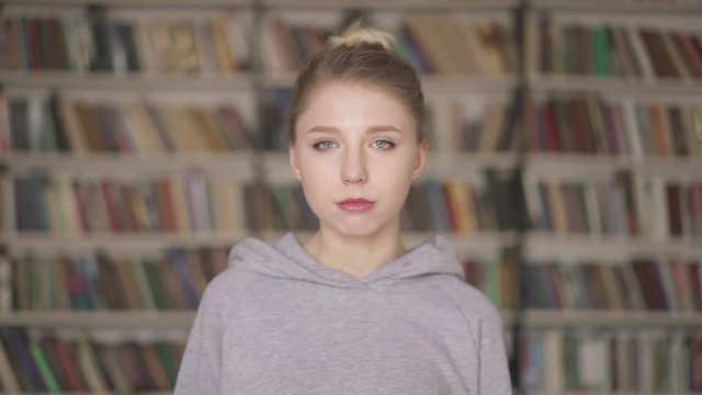 Portrait of smart and confident beautiful female blonde student standing in library. Young girl green eyes in grey hoodie standing on the background of bookshelves in the library.