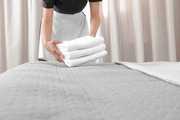 Young maid putting stack of fresh towels on bed in hotel room, closeup