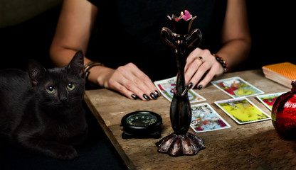 The fortune teller lays out on a wooden table the tarot cards by the light of a candle. Black cat sitting near the table. Selective focus. - obrazy, fototapety, plakaty