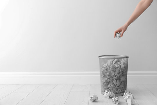 Woman throwing crumpled paper into metal bin against light wall. Space for text
