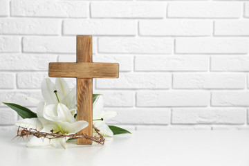 Wooden cross, crown of thorns and blossom lilies on table against brick wall, space for text