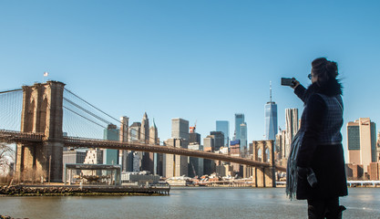 Young Woman tourist by the river in Dumbo taking pictures of Brooklyn Bridge and Cityscape of New...
