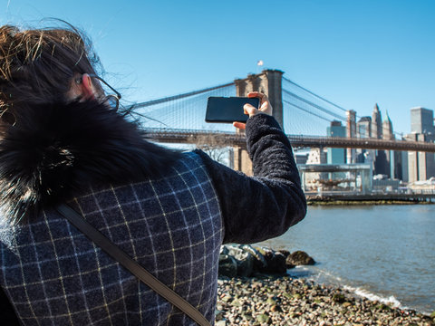 Young Woman tourist by the river in Dumbo taking pictures of Brooklyn Bridge and Cityscape of New York skyline.