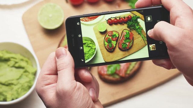 A man photographs on a smartphone sandwiches with avocados, cherry tomatoes and salmon fillets.