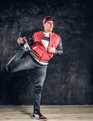 Professional breakdancer wearing fashionable clothes, warming up and stretching his legs for performance. Studio photo against a dark textured wall