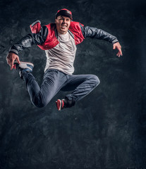 Fototapeta na wymiar Emotional stylish dressed guy performing break dance jumping. Studio photo against a dark textured wall