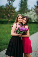Two sisters walking outside in spring park. They hold a bouqet of lilac and smile