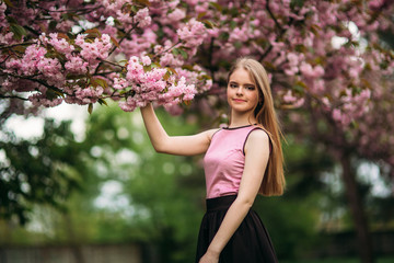 Portrait of European female near the Japanese flowering tree of sakura. Blond hair girl outside