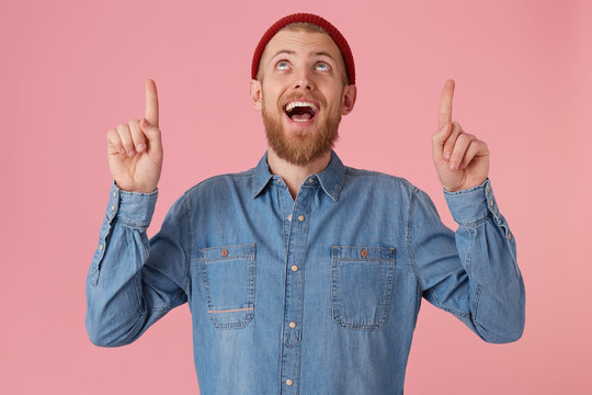 Emotive pleased young bearded guy looks up with excitement and toothy smile, keeps hands up and pointing with fore fingers upwards, dressed in a denim shirt, isolated on a pink background