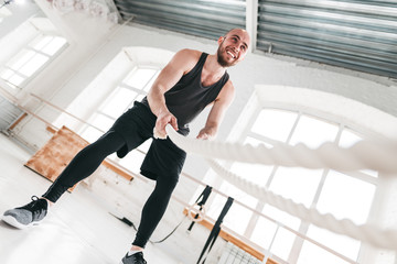 Smiling man workout with battle ropes at light gym. Muscular sportsman doing crossfit excursion...