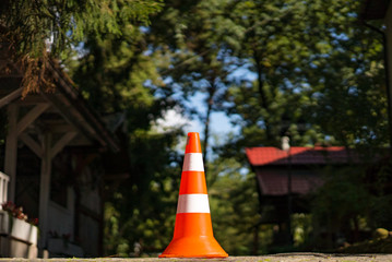 Street cone of two colors with orange and white standing over the natural background. Warning striped plastic orange parking cone near the houses in forest.
