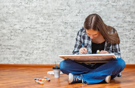 Portrait Of Young Teenager Brunette Girl With Long Hair Sitting On Floor And Drawing Picture On Gray Wall Background With Copy Space