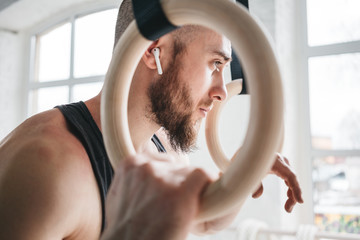 Male bearded athlete with wireless headphones resting and holding gymnastics rings at light gym....